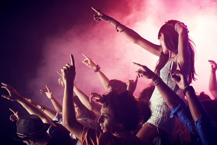 A young woman with long dark hair wearing a white minidress, sitting on someone's shoulders at a gig, she and everyone in the audience have their arms in the air. 